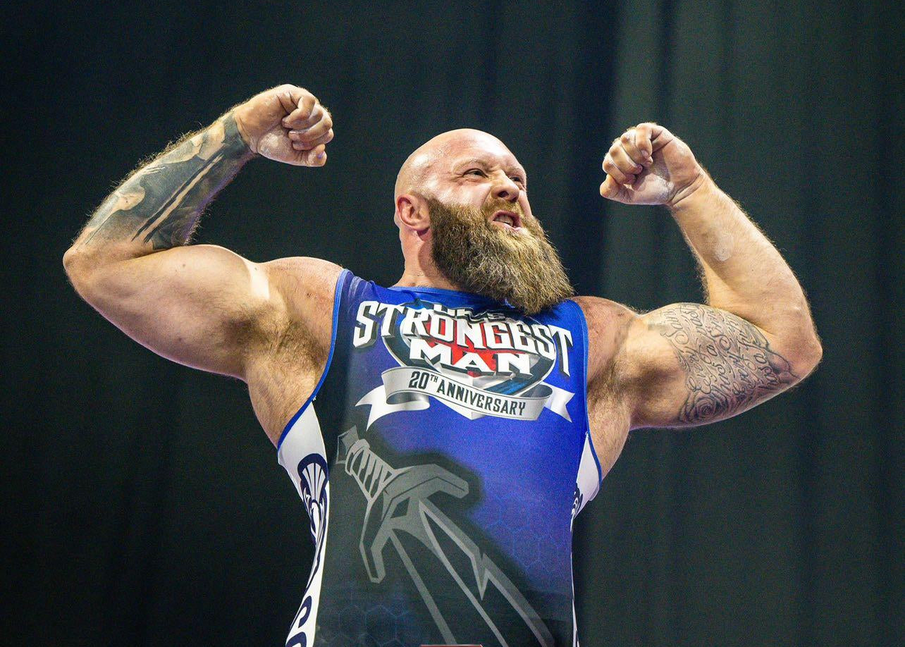 A muscular bearded man flexing his biceps while wearing a blue and white singlet that reads 'STRONGEST MAN 20th Anniversary' and 'The Power of Strongman'.